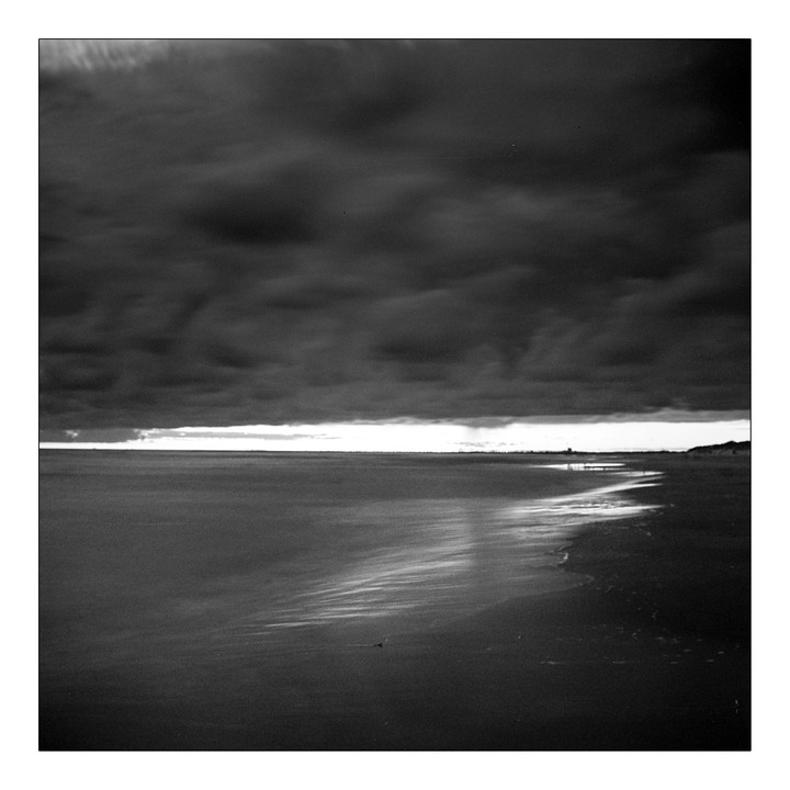 A dark, cloudy sky hangs over a beach at night, with a bright horizon line and waves washing onto the shore.