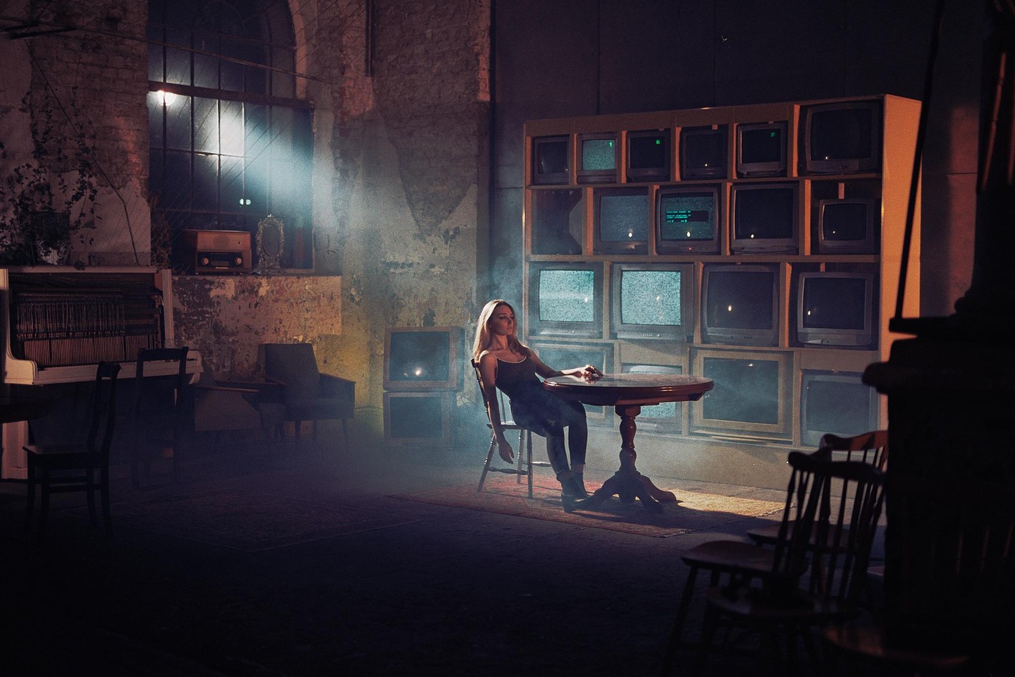 A woman sits at a table in a dimly lit room with a wall of televisions behind her.