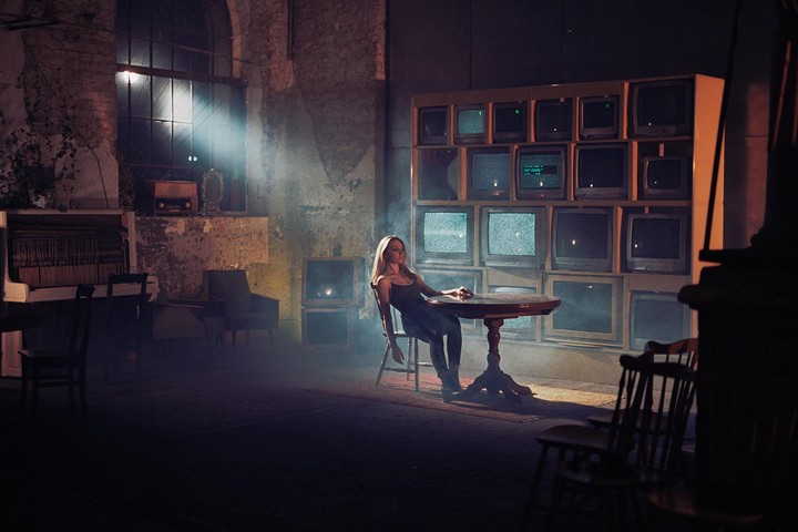 A woman sits at a table in a dimly lit room with a wall of televisions behind her.