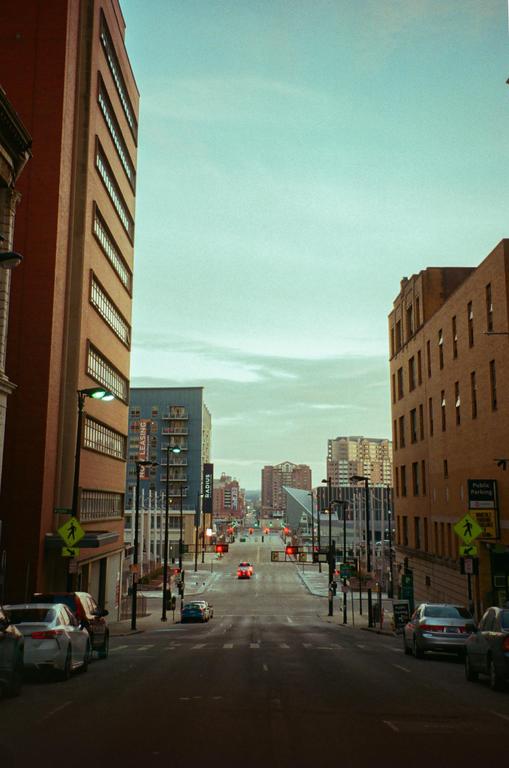 A wide street with parked cars on both sides leads down a hill towards distant buildings under a pale sky.