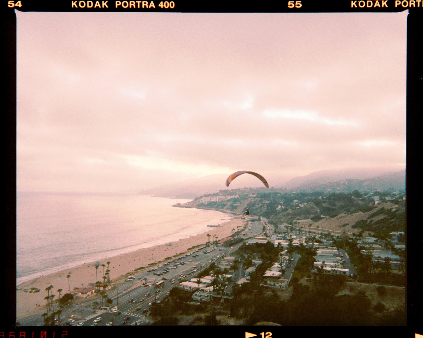 A paraglider soars above a coastal town with a beach and highway.