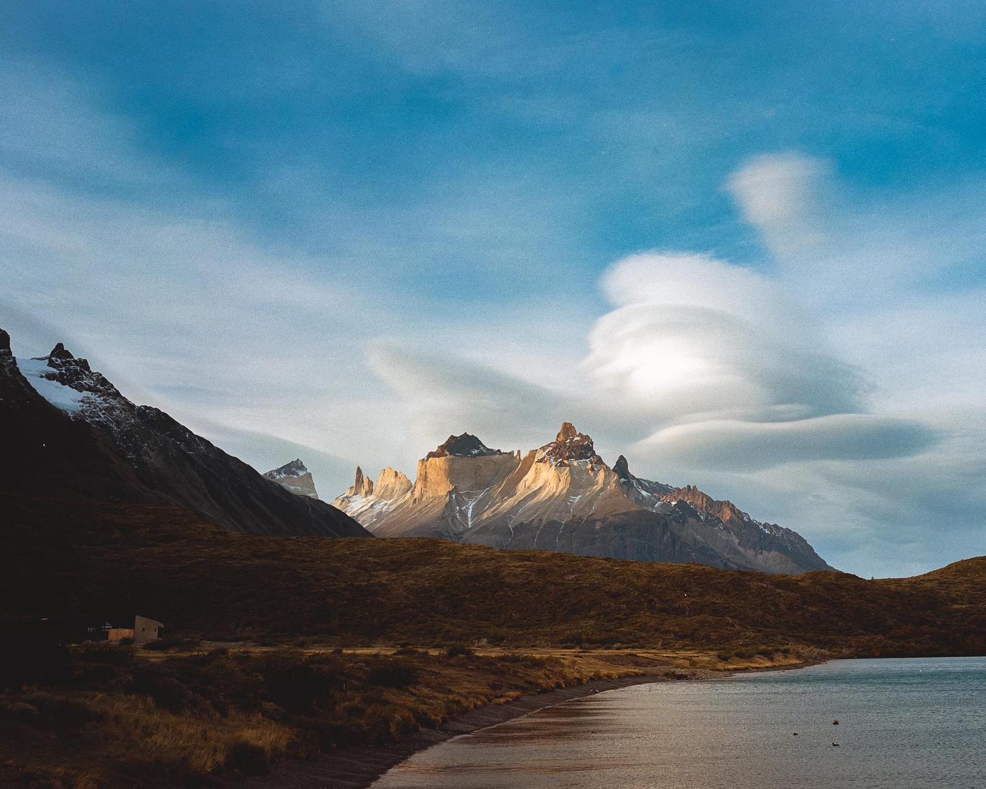 A body of water with a small building on the shore is in the foreground of a mountain range under a cloudy sky.