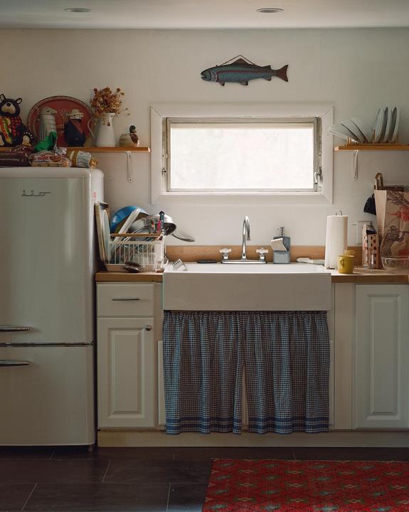 A cozy kitchen features a white refrigerator, a sink with a checkered curtain, and a decorative fish hanging above a window.