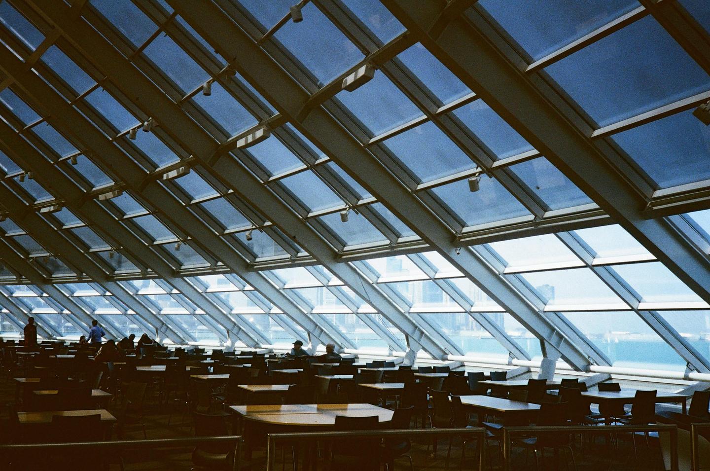A large room with many tables and chairs is illuminated by natural light coming through a glass ceiling and wall.