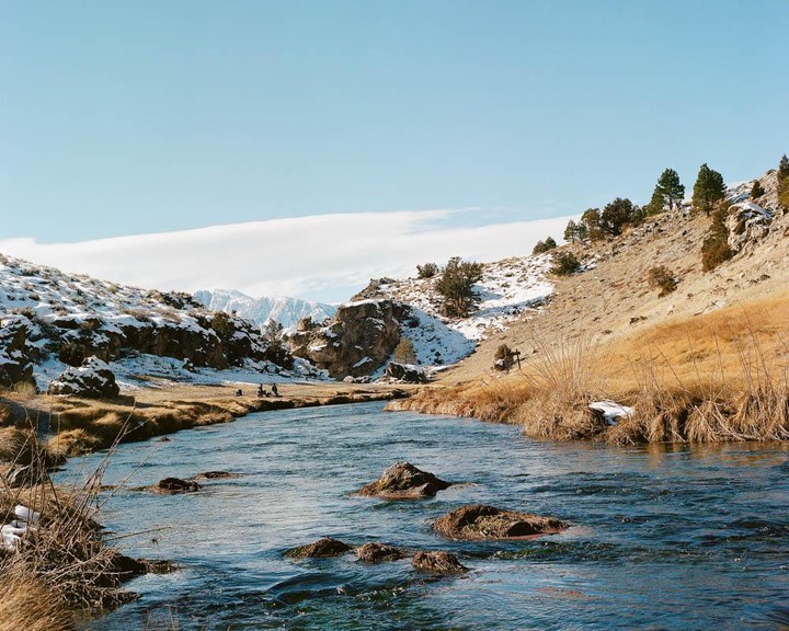 A river flows through a snowy and grassy landscape with mountains in the background.