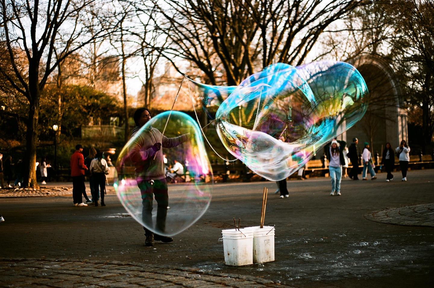 A man creates large, colorful bubbles in a park with bare trees and other people in the background.