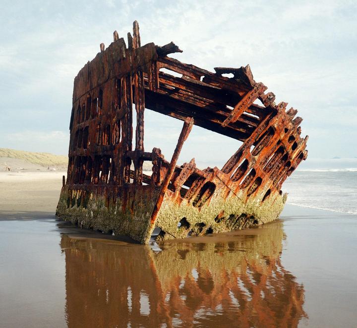 The rusted remains of a shipwreck stand on a sandy beach with waves crashing nearby.