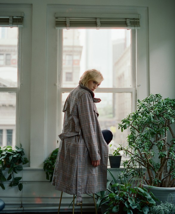 A woman in a plaid coat stands in front of a window with several plants around her.