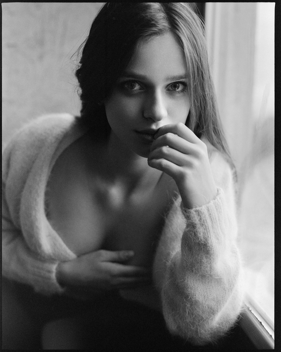 A woman with long hair and a fuzzy sweater looks at the viewer, with one hand near her mouth and the other covering her chest.
