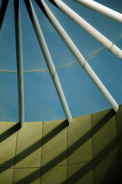 Sunlight casts shadows from white beams onto a curved wall under a blue sky.