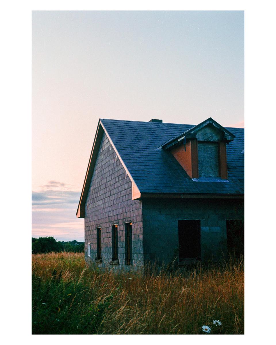 An old house with a dark roof stands in a field of tall grass under a pale sky.