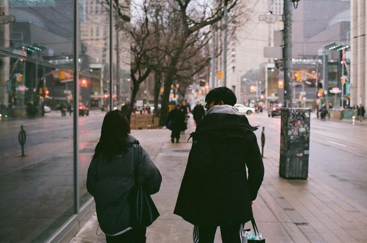 Two people walk along a city street with buildings and trees in the background.