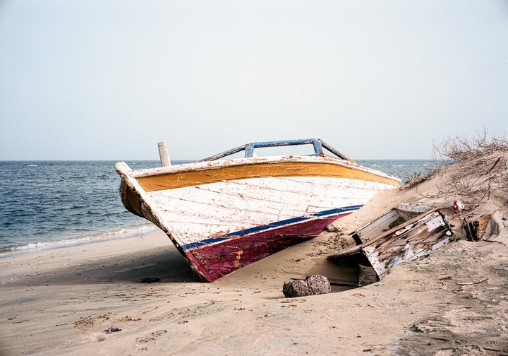 A weathered boat with peeling paint rests on a sandy beach next to the ocean.