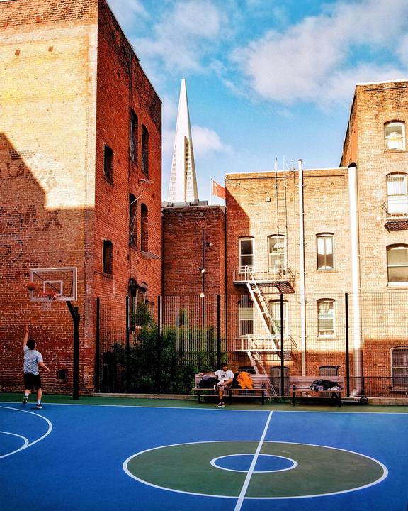 A man shoots a basketball on a blue court surrounded by brick buildings, with a tall white building visible in the background.