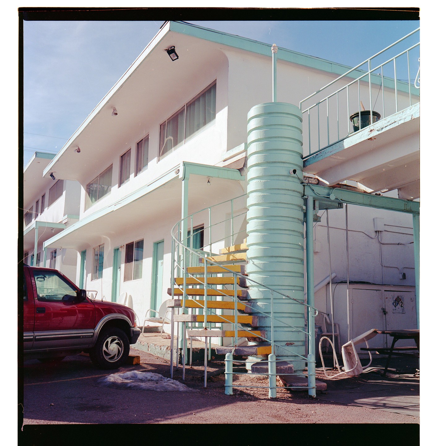 A red truck is parked in front of a white motel with a turquoise spiral staircase.