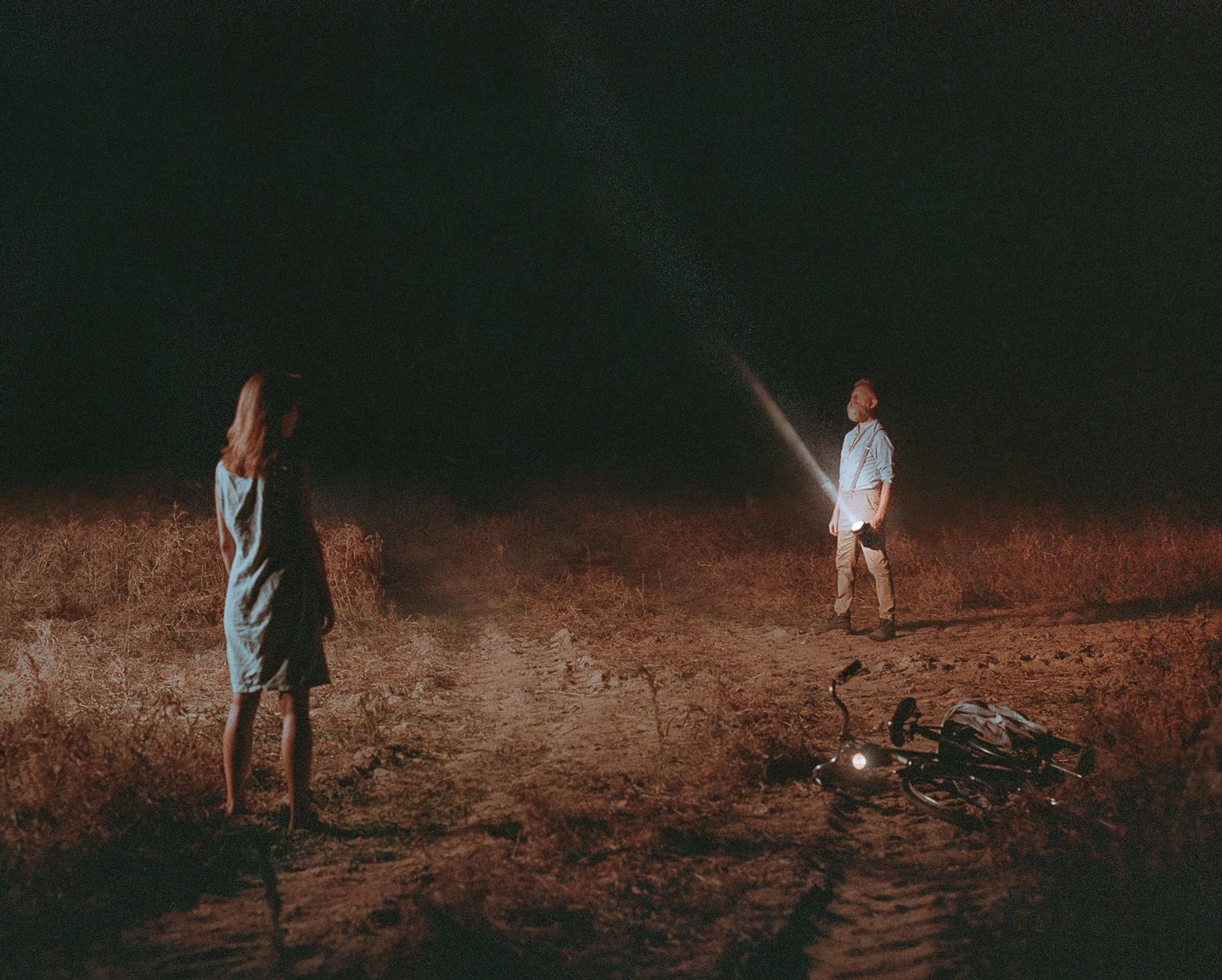 A man shines a flashlight at a woman in a field at night, with a bicycle lying on the ground.