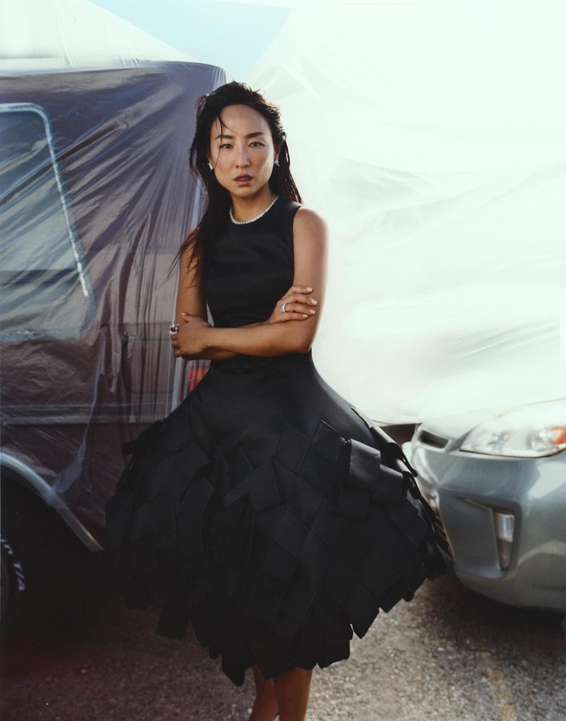 A woman in a black dress stands with her arms crossed in front of two vehicles covered in plastic sheeting.