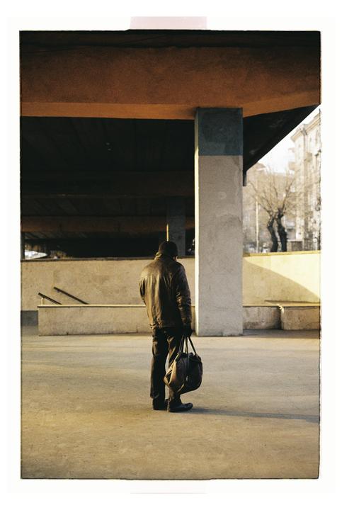 A man in a leather jacket and hat stands with a bag under a concrete overhang.