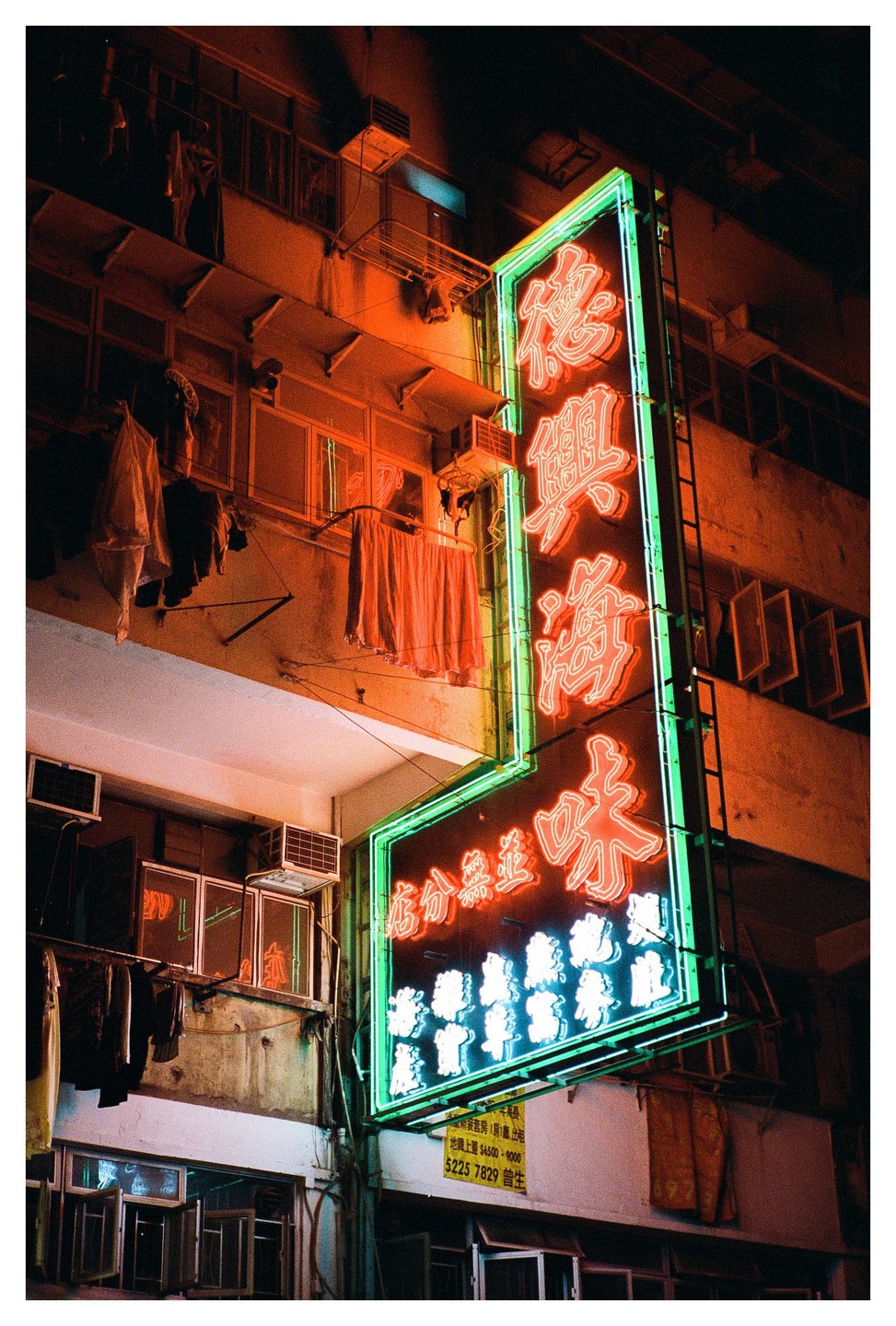 A large neon sign with Chinese characters illuminates the side of a building where laundry hangs to dry.