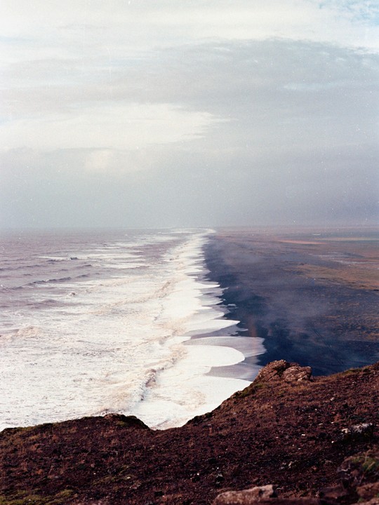 A long stretch of black sand beach with white waves crashing on the shore is seen from a high vantage point.