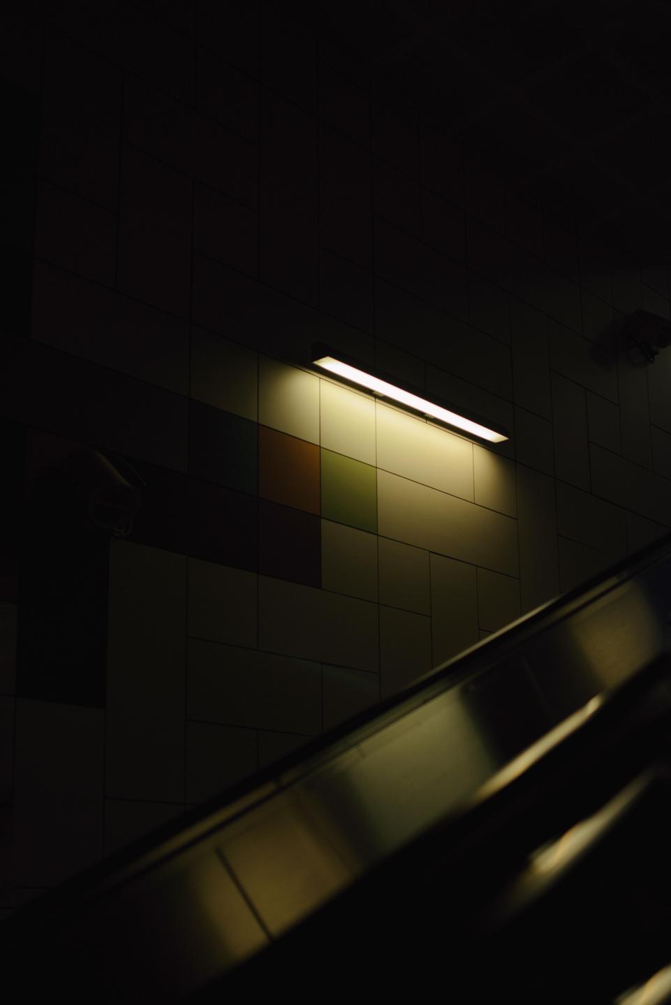 A fluorescent light illuminates a tiled wall above an escalator.