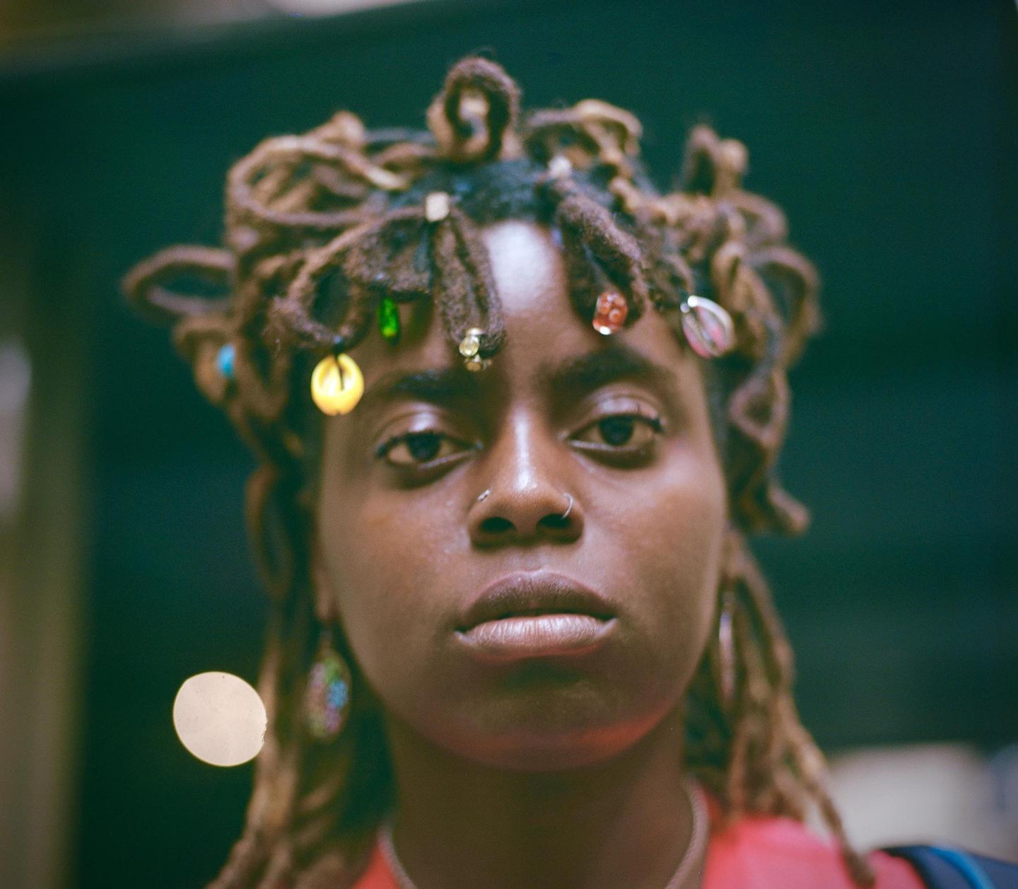 A woman with dreadlocks adorned with beads and shells looks directly at the viewer.