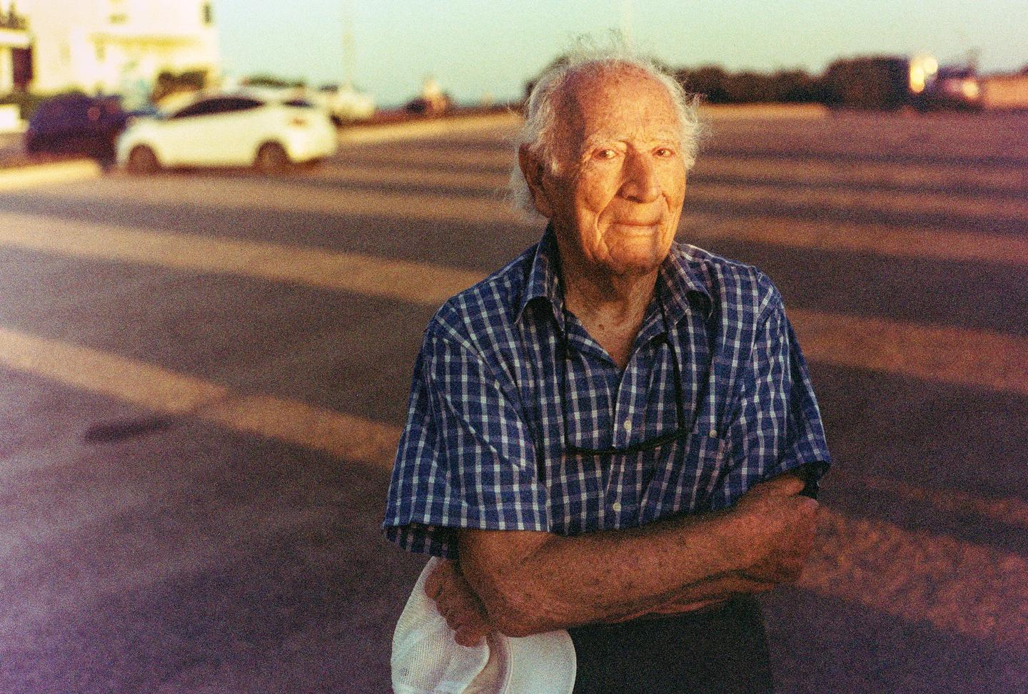 An elderly man in a plaid shirt stands with his arms crossed, holding a white hat, with cars and a street in the background.