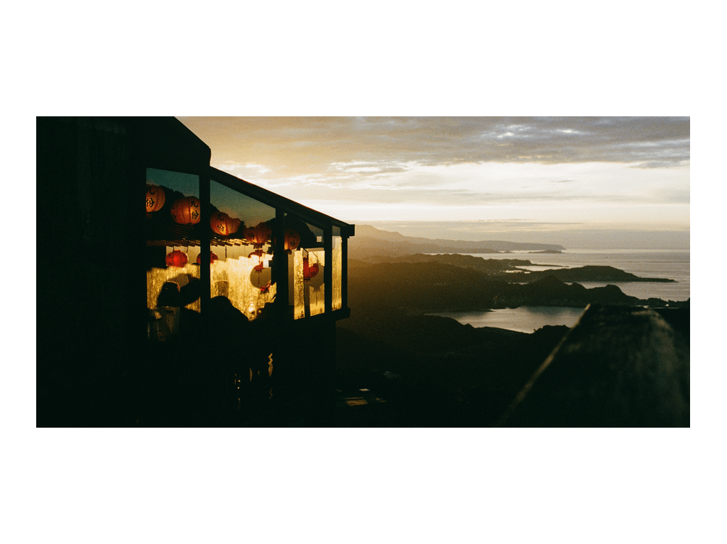 A building with illuminated lanterns is silhouetted against a sunset over a bay with mountains in the distance.
