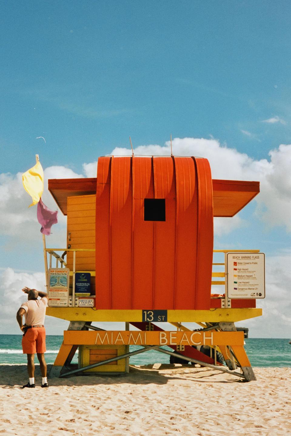 A man stands on a sandy beach looking out at the ocean next to a colorful lifeguard stand.