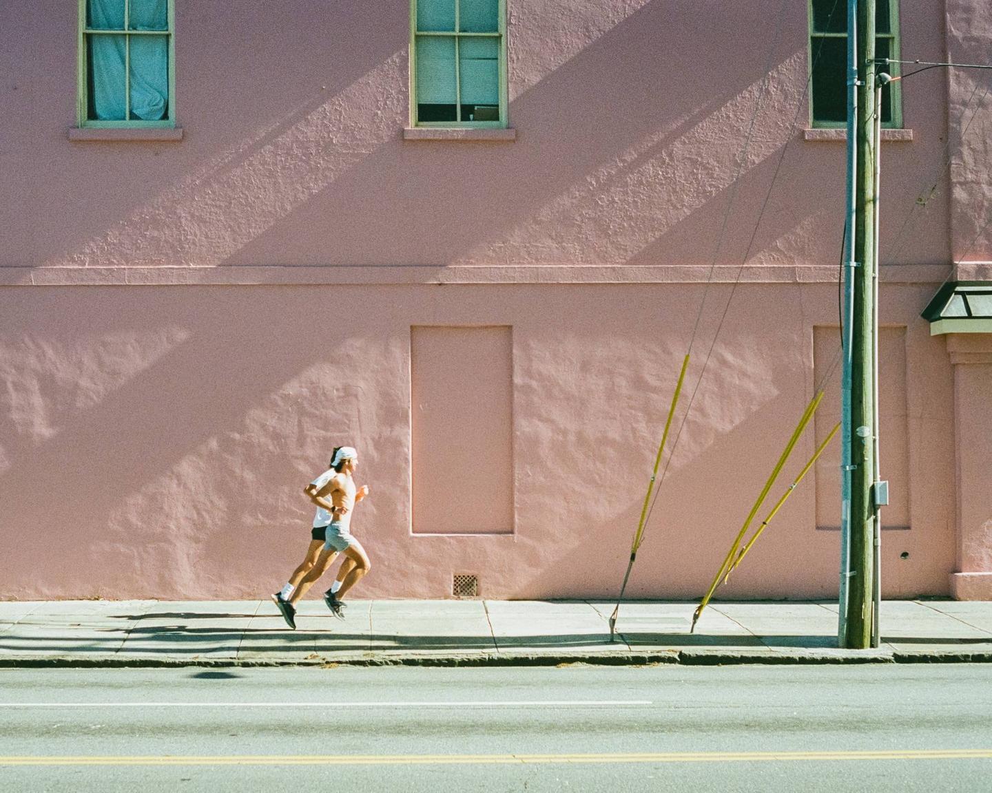 Two men jog along a sidewalk next to a pink building with windows and a utility pole.