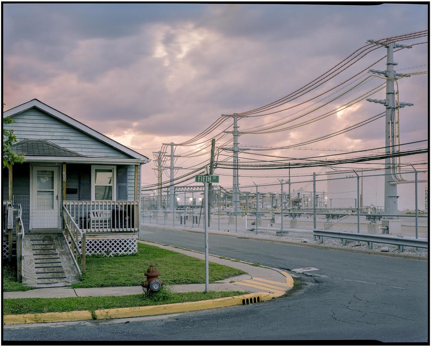 A small house with a porch sits next to a street with a fire hydrant and a power station under a cloudy sky.