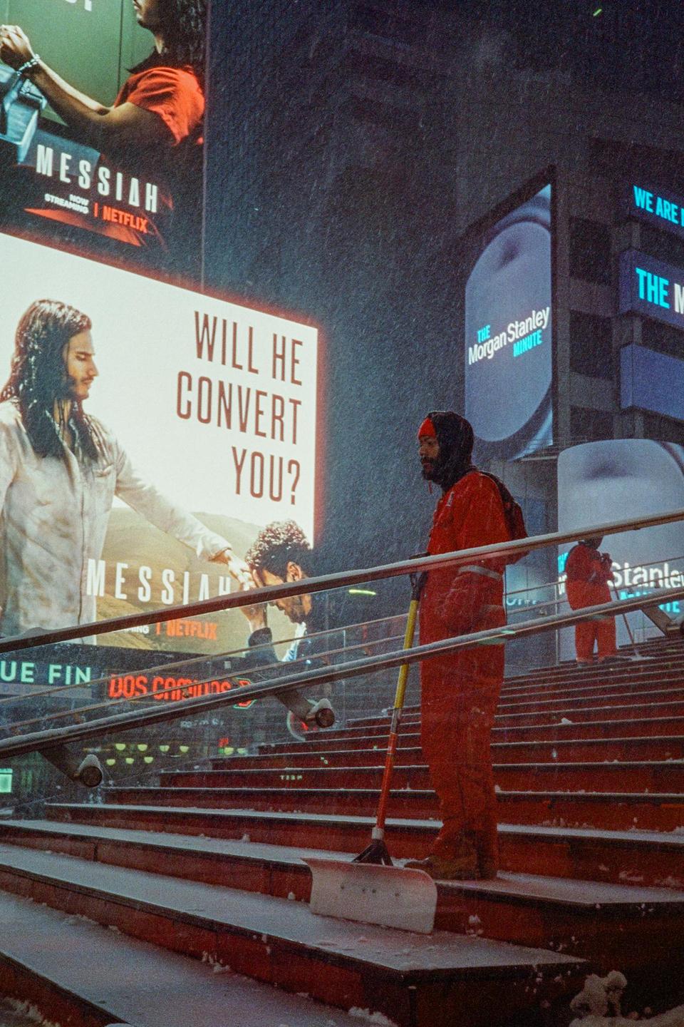 A man in a red jumpsuit stands on a snowy staircase next to a snow shovel, with bright billboards in the background.