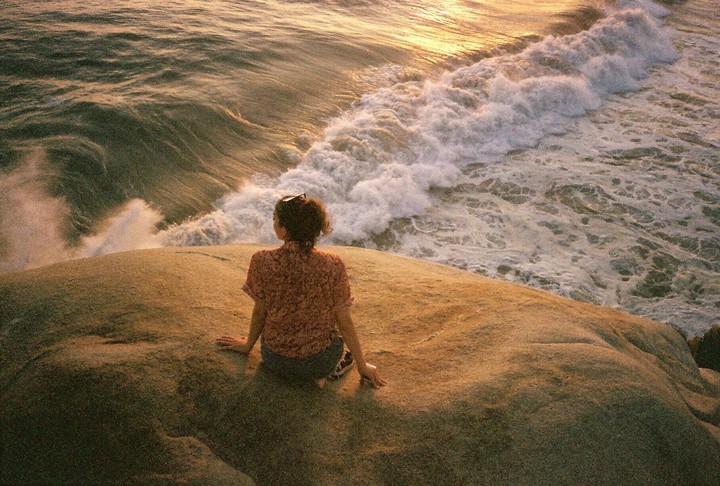 A woman sits on a rock overlooking the ocean as waves crash on the shore during golden hour.