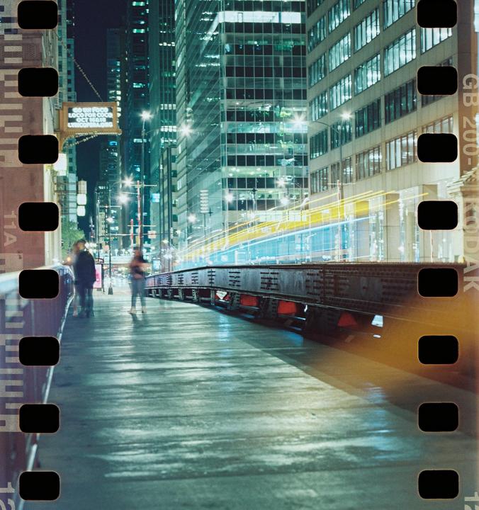 Two people stand on a bridge next to a blurred train with city buildings in the background at night.