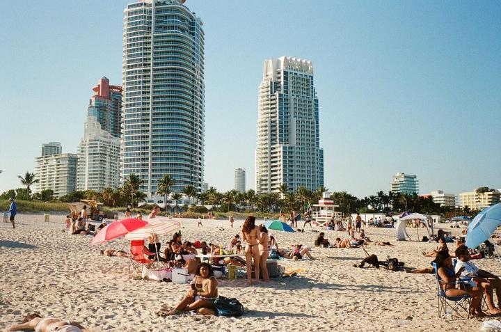 A wide shot shows a crowded beach with people sunbathing and swimming, with tall buildings and palm trees in the background.
