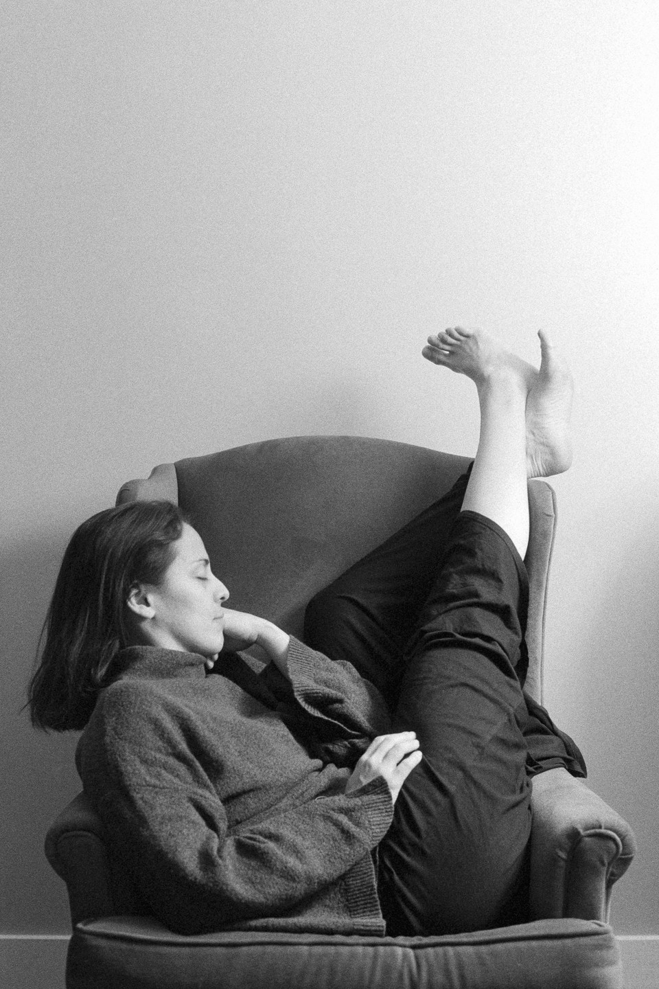A woman reclines in an armchair with her legs propped up against a wall, her eyes closed.