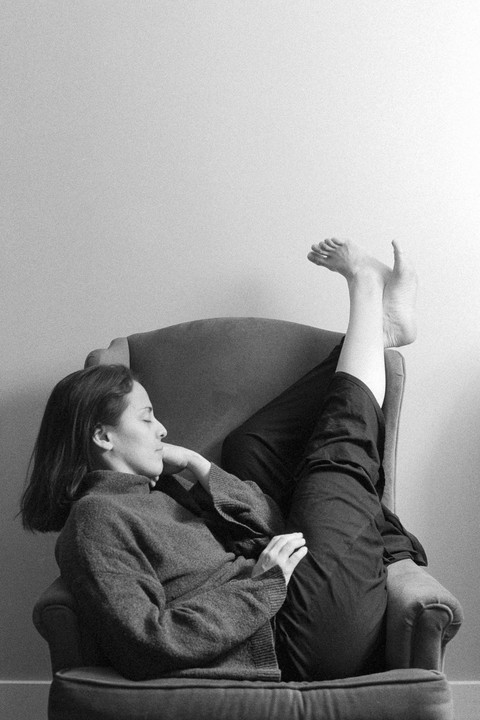 A woman reclines in an armchair with her legs propped up against a wall, her eyes closed.