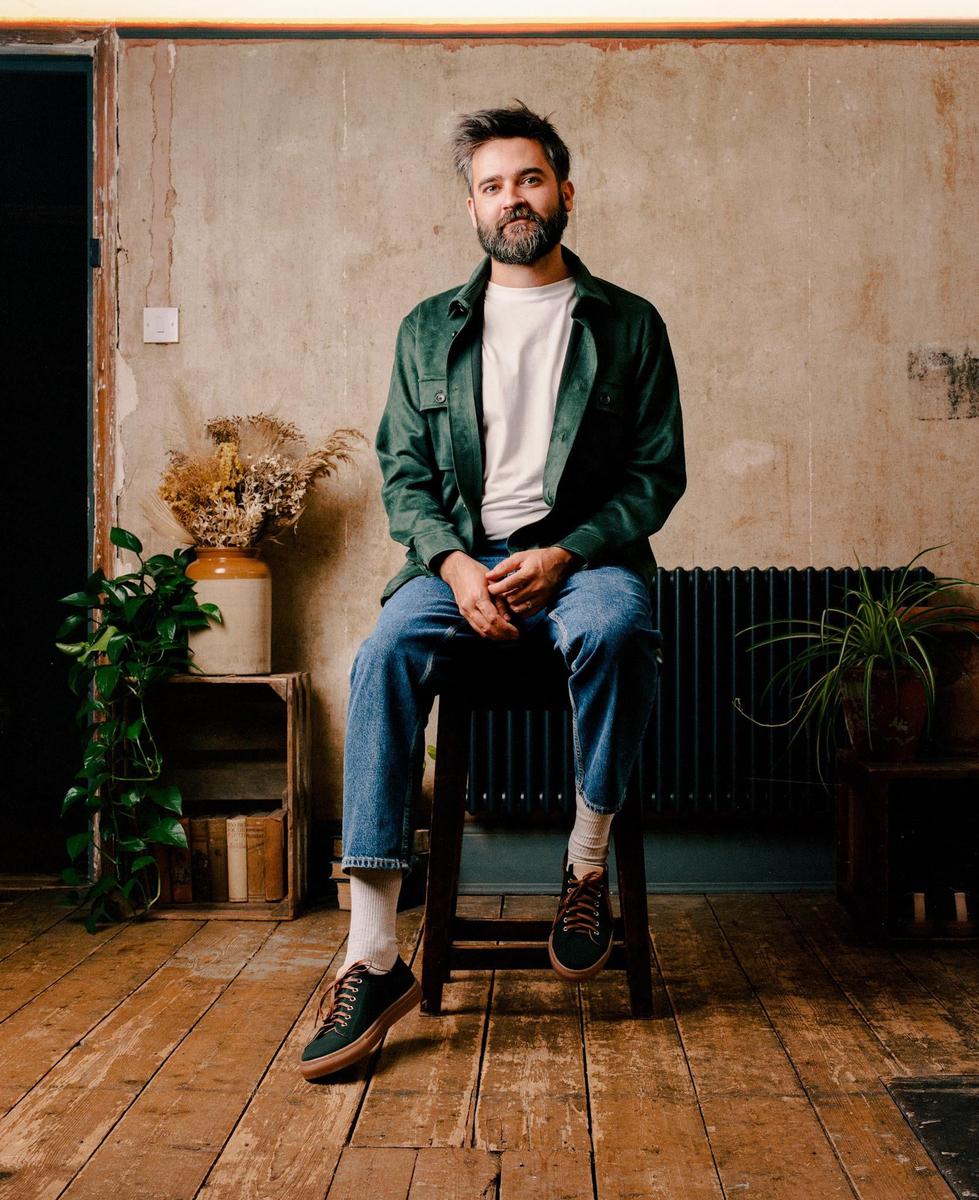 A man with a beard sits on a stool in a room with wooden floors and a distressed wall.