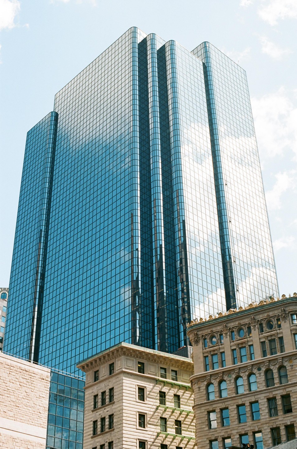 A tall glass skyscraper reflects the sky and clouds, towering over older, ornate buildings.