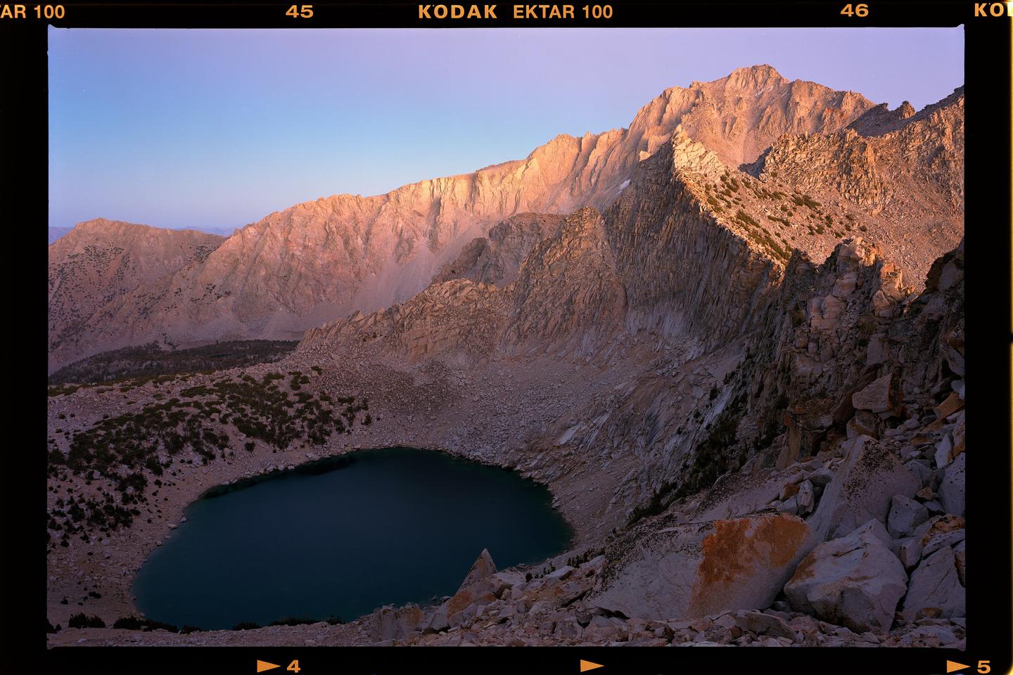 A high-angle landscape view shows a mountain lake surrounded by rocky slopes and peaks under a clear sky.