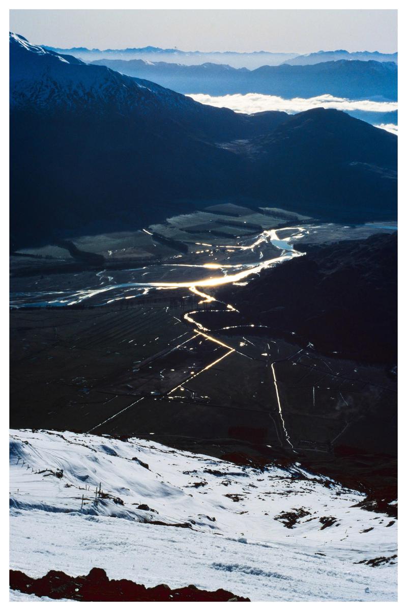 A winding river reflects sunlight through a valley surrounded by mountains, with a snowy slope in the foreground.