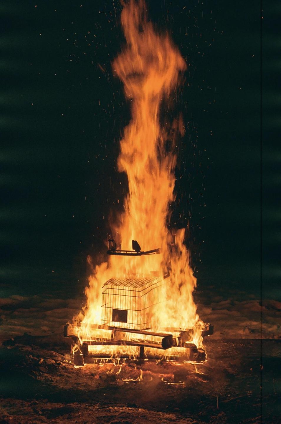 A tall bonfire burns brightly in the dark, with a metal cage visible within the flames.