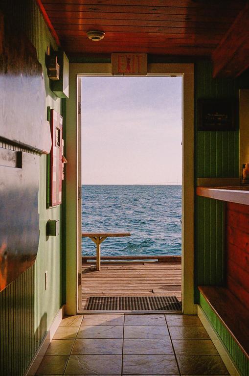 A view from inside a building shows a wooden pier extending into the ocean under a bright sky.