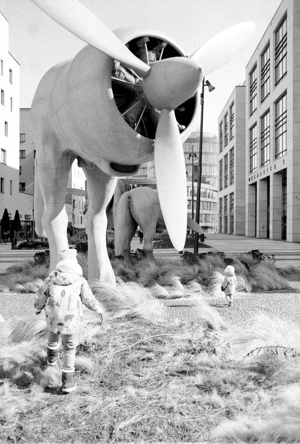 Two children walk through tall grass towards large elephant sculptures with airplane propellers for heads in a city square.
