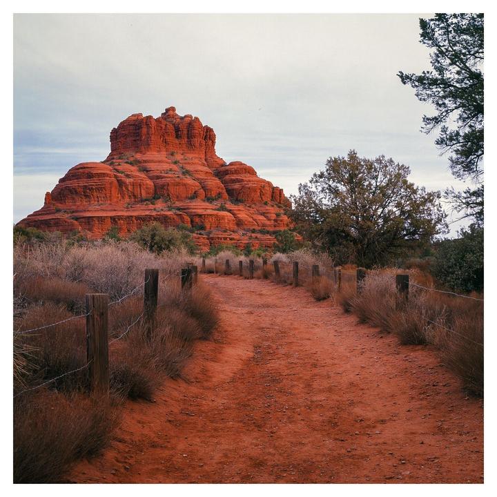 A red dirt path with a barbed wire fence leads towards a large red rock formation under a cloudy sky.
