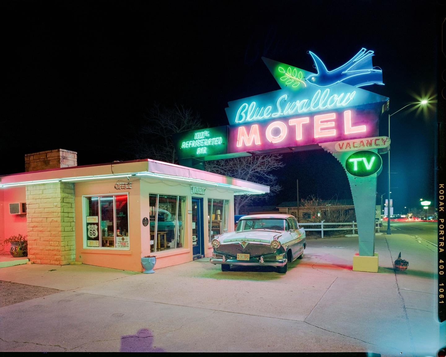 A vintage car is parked in front of the Blue Swallow Motel, which is illuminated by neon lights at night.