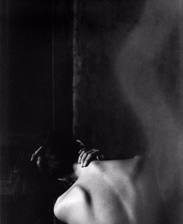 A woman with her back to the viewer is seen from the shoulders up, with her hands on her head and neck.