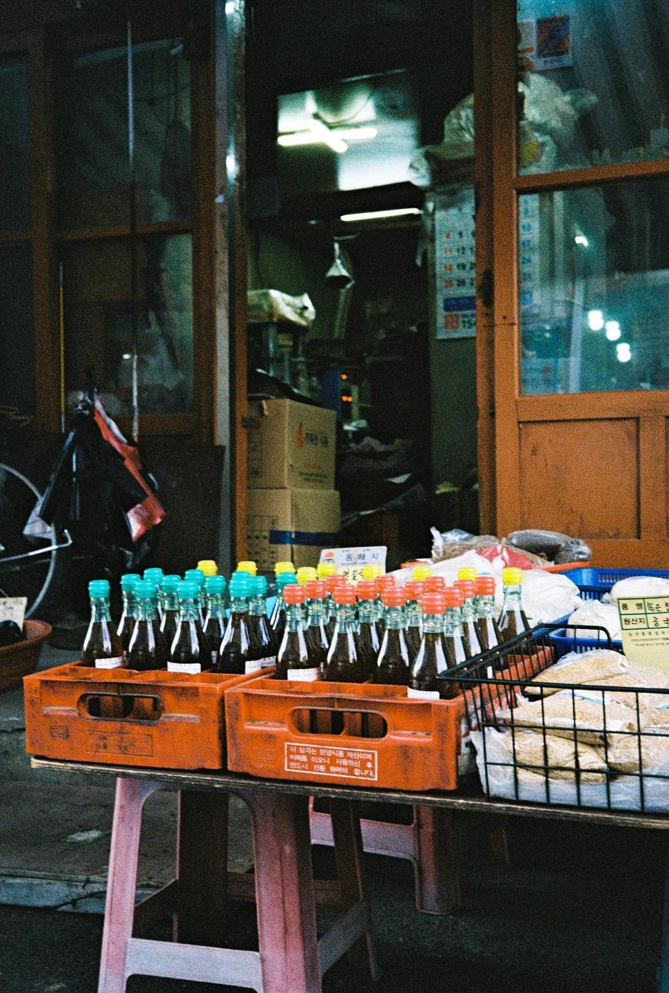 Bottles of sauce with colorful caps sit in orange crates on a table outside a store.