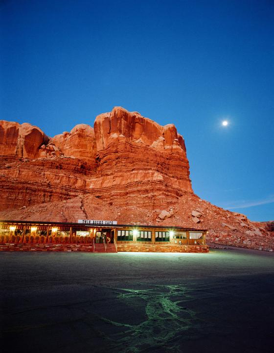A cafe with illuminated windows sits at the base of a large red rock formation under a blue sky with a full moon.