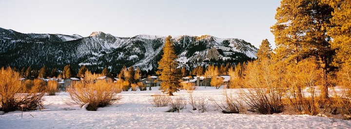 A snowy landscape with mountains, evergreen trees, and houses is illuminated by golden light.